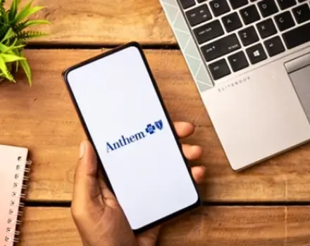 Hand holding smartphone displaying Anthem insurance company logo on screen next to laptop on wooden desk, representing digital access to healthcare insurance benefits and coverage information
