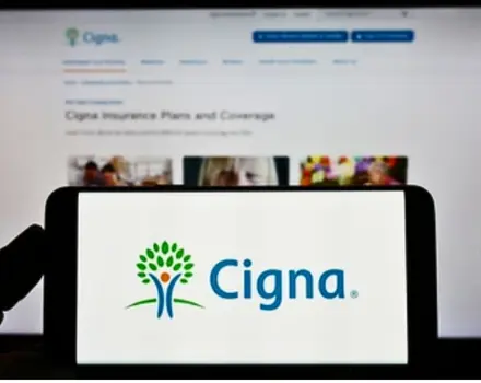 Hands holding tablet displaying Cigna insurance company logo with tree symbol on screen, with insurance plan information visible in background, representing digital access to healthcare coverage and benefits
