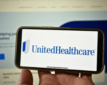 Hand holding smartphone displaying UnitedHealthcare insurance company logo on screen with insurance documents visible in background, representing digital access to healthcare coverage and benefits information