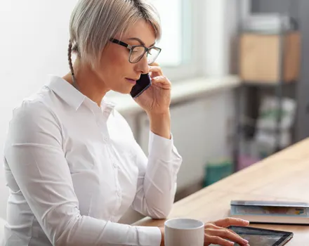 Professional woman with glasses and blonde hair speaking on phone while working at desk, representing consultation and support services for mental health care