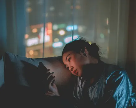 Person lying in dim lighting looking at phone screen with city lights blurred in background, representing someone privately taking an online depression assessment during a difficult moment