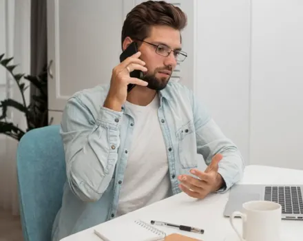 Professional man in glasses speaking on phone while sitting at desk with laptop, representing connection with mental health professionals and accessible therapy services