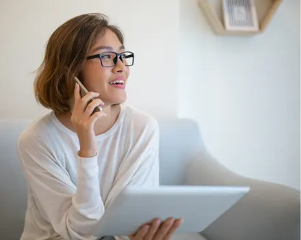 Woman with glasses smiling while talking on phone and holding tablet, representing positive interaction with mental health support services and professional consultation