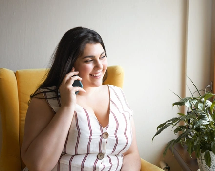 Smiling woman in striped top talking on phone while sitting in bright room with yellow chair, illustrating positive engagement with mental health support services and professional consultation